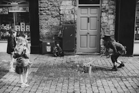 A street performer wearing a top hat and casual clothes is interacting with a group of children. The performer is squatting and using a tool to handle a bucket placed on a cobblestone street. The scene is set in front of a brick wall and a closed door, creating an urban backdrop.
