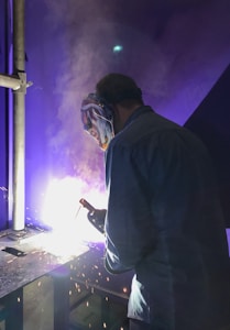 A person wearing a protective mask and a jacket is engaged in welding. Bright sparks and a vivid glow illuminate the work area, creating a dramatic contrast against the dark environment.