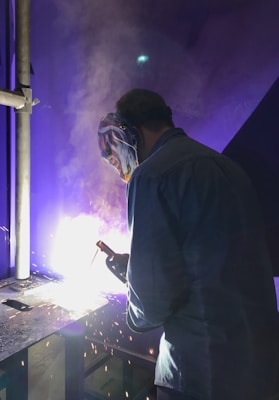 A person wearing a protective mask and a jacket is engaged in welding. Bright sparks and a vivid glow illuminate the work area, creating a dramatic contrast against the dark environment.