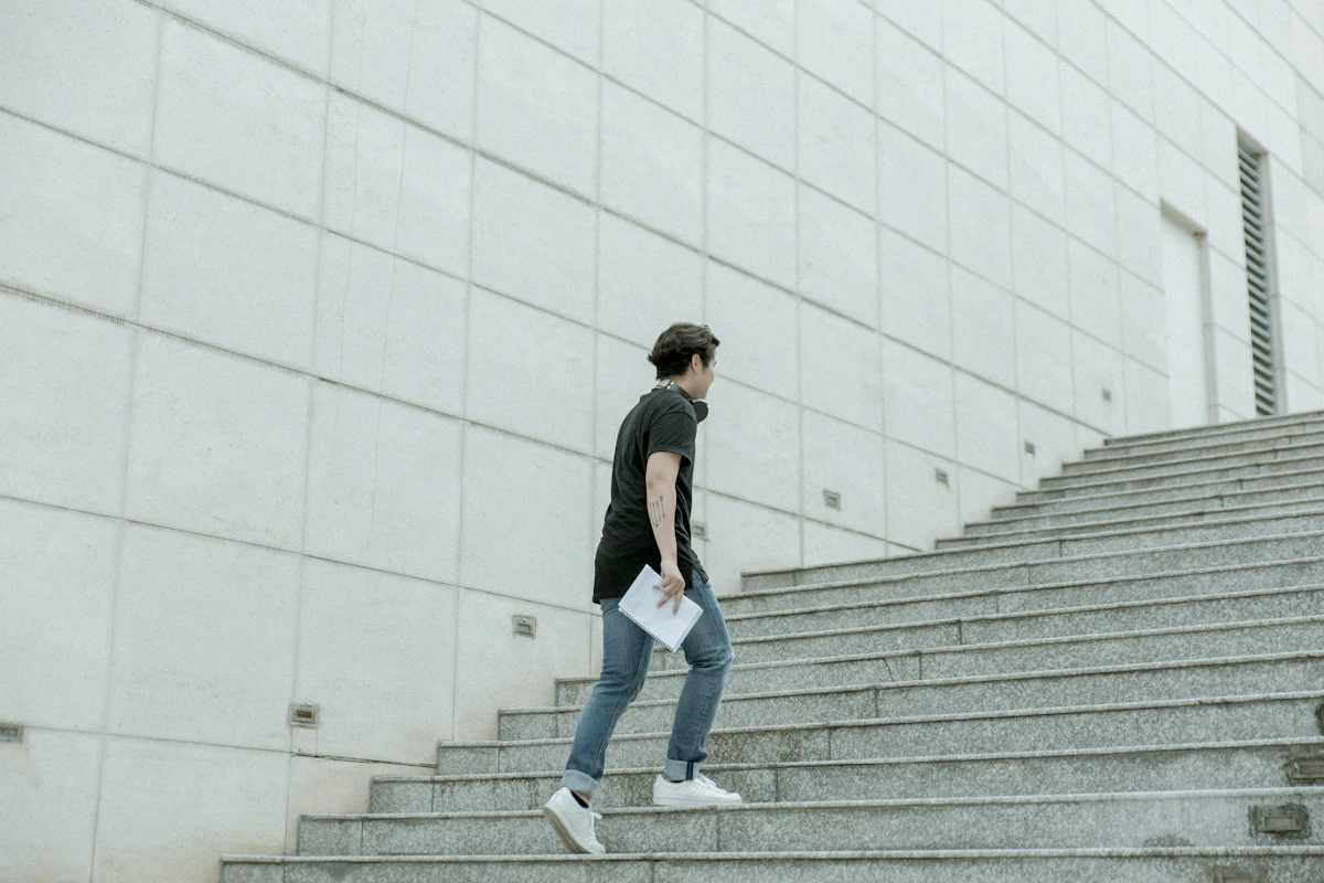A young man climbs a wide grey staircase beside a pale wall, holding papers