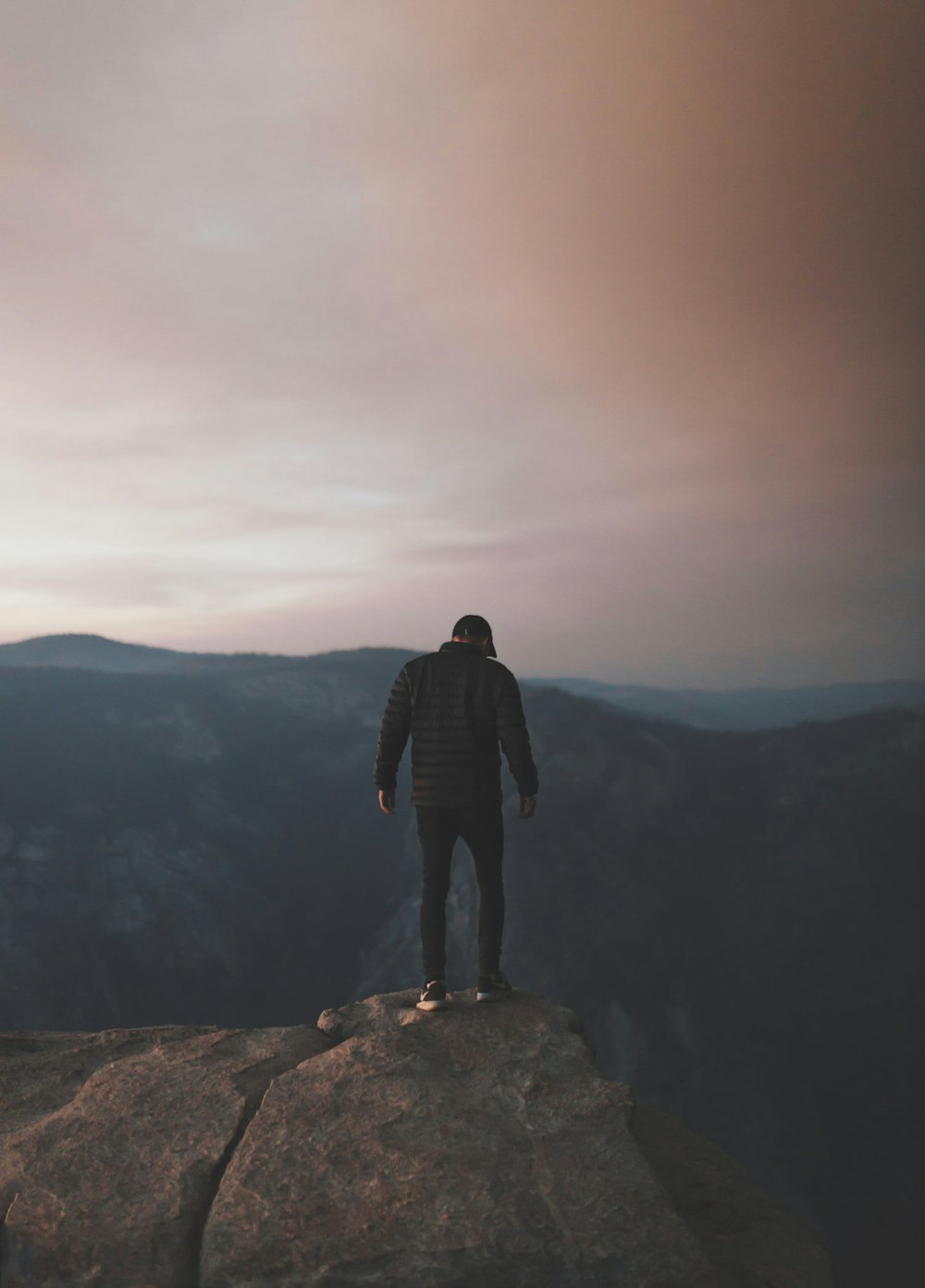 Uomo che indossa una giacca a bolle nera sulla collina rocciosa della montagna di fronte alle montagne sotto cieli nuvolosi bianchi, rosa e blu durante il giorno