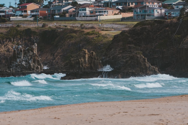 Waves crash against rocky cliffs, with a picturesque view of houses perched on a hillside in the background. The beach in the foreground is sandy, while the ocean displays various shades of blue, showcasing a dynamic coastal scene.