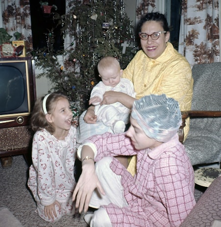 A family scene with a woman holding a baby, a young girl in pajamas laughing, and another person seated on the floor. A small Christmas tree is decorated with lights and tinsel in the background. A vintage television is placed in the corner.