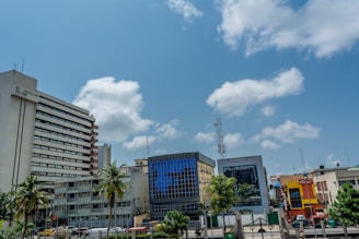 A cityscape featuring several modern buildings, including a tall white building labeled 'St. Nicholas House' on the left and a blue-glass building in the center. Palm trees and other greenery are visible among the urban infrastructure. The sky is a clear blue with scattered white clouds.