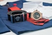 A close-up of fashion accessories including a black belt with a silver Medusa logo buckle, a rose gold analog watch with a black strap, and a folded red tie on top of blue and white clothing material.