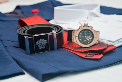 A close-up of fashion accessories including a black belt with a silver Medusa logo buckle, a rose gold analog watch with a black strap, and a folded red tie on top of blue and white clothing material.