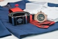 A close-up of fashion accessories including a black belt with a silver Medusa logo buckle, a rose gold analog watch with a black strap, and a folded red tie on top of blue and white clothing material.