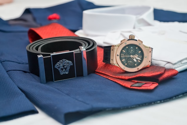 A close-up of fashion accessories including a black belt with a silver Medusa logo buckle, a rose gold analog watch with a black strap, and a folded red tie on top of blue and white clothing material.