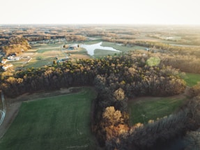 An aerial view of a rural landscape featuring expansive fields, a dense woodland area, and a small body of water. Several houses and farm buildings are scattered throughout the fields, with a network of roads connecting them. The scene is bathed in the warm light of either sunrise or sunset.