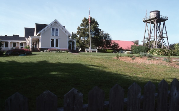 A picturesque farmhouse with white siding and a gabled roof is surrounded by lush greenery and a well-maintained lawn. A large red barn is visible in the background, and a wooden water tower stands to the right. The scene includes a flagpole with the American flag and a variety of shrubs and flowers in bloom.