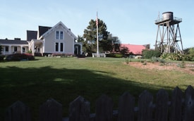 A picturesque farmhouse with white siding and a gabled roof is surrounded by lush greenery and a well-maintained lawn. A large red barn is visible in the background, and a wooden water tower stands to the right. The scene includes a flagpole with the American flag and a variety of shrubs and flowers in bloom.