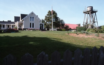 A picturesque farmhouse with white siding and a gabled roof is surrounded by lush greenery and a well-maintained lawn. A large red barn is visible in the background, and a wooden water tower stands to the right. The scene includes a flagpole with the American flag and a variety of shrubs and flowers in bloom.
