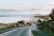 A coastal road winds down towards a sandy beach with the sea in the background. Small houses with chimneys line the street, and several vehicles are parked along the road. The landscape is slightly hilly, with green bushes and grass along the sides. There is a castle-like structure visible in the distance on the land jutting into the sea.