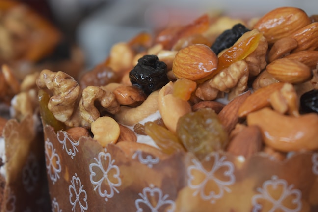 A close-up of a richly decorated baked pastry topped with a mix of nuts and dried fruits. The dough is wrapped in a brown paper liner with white floral patterns. Walnuts, almonds, cashews, raisins, and candied fruits are visible, creating a textured and colorful topping.