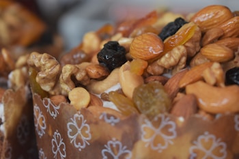 A close-up of a richly decorated baked pastry topped with a mix of nuts and dried fruits. The dough is wrapped in a brown paper liner with white floral patterns. Walnuts, almonds, cashews, raisins, and candied fruits are visible, creating a textured and colorful topping.