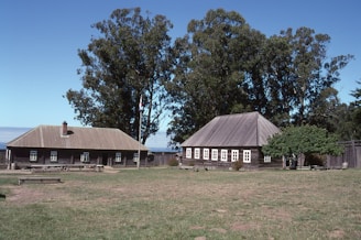 Two rustic wooden buildings with pitched roofs sit on a grassy field. Large trees provide a backdrop, and a flagpole is positioned between the buildings. Several picnic tables are scattered across the lawn.