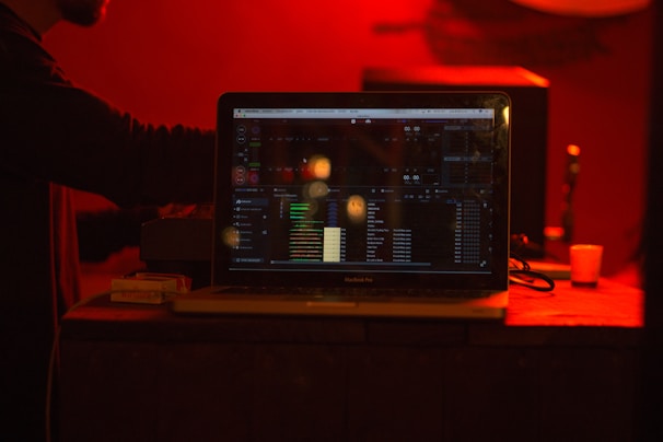 A laptop displaying music or audio software with colorful interface elements sits on a table in a dimly lit room with a deep red ambiance. In the background, there is a hint of a person interacting with equipment, and various objects like a small glass and a pack of cigarettes are on the table.