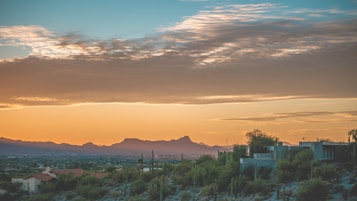 The image captures a serene desert landscape during sunset with a clear view of mountains in the distance. The sky is a gradient of warm orange and soft blue hues, dotted with scattered clouds. Cacti and desert shrubs are visible in the foreground, while a few houses are nestled amidst the natural scenery.