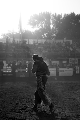 A lone cowboy dressed in traditional rodeo attire walks on a dirt arena. The sun is setting or rising, creating a bright glare and casting long shadows. In the background, there are bleachers filled with spectators, partially obscured by the glow of sunlight.