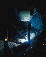 A welder wearing a protective helmet is engaged in welding metal pieces together. The scene is dimly lit, with bright sparks and blue light illuminating the area around the welding point. The details on the helmet are slightly visible due to the light reflections.