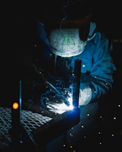 A welder wearing a protective helmet is engaged in welding metal pieces together. The scene is dimly lit, with bright sparks and blue light illuminating the area around the welding point. The details on the helmet are slightly visible due to the light reflections.