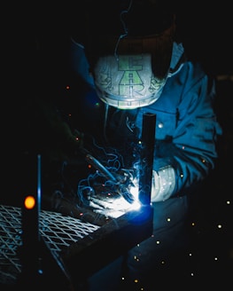 A welder wearing a protective helmet is engaged in welding metal pieces together. The scene is dimly lit, with bright sparks and blue light illuminating the area around the welding point. The details on the helmet are slightly visible due to the light reflections.