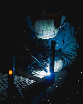 A welder wearing a protective helmet is engaged in welding metal pieces together. The scene is dimly lit, with bright sparks and blue light illuminating the area around the welding point. The details on the helmet are slightly visible due to the light reflections.
