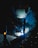 A welder wearing a protective helmet is engaged in welding metal pieces together. The scene is dimly lit, with bright sparks and blue light illuminating the area around the welding point. The details on the helmet are slightly visible due to the light reflections.