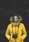A person is wearing a bright yellow, puffy jacket with a fur-lined hood. They have a camera hanging from their neck, displaying the Nikon brand. The background is composed of dark greenery, possibly coniferous trees, adding a natural and slightly moody atmosphere to the scene. The individual has their hands in their jacket pockets, and their expression seems calm.
