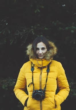 A person is wearing a bright yellow, puffy jacket with a fur-lined hood. They have a camera hanging from their neck, displaying the Nikon brand. The background is composed of dark greenery, possibly coniferous trees, adding a natural and slightly moody atmosphere to the scene. The individual has their hands in their jacket pockets, and their expression seems calm.