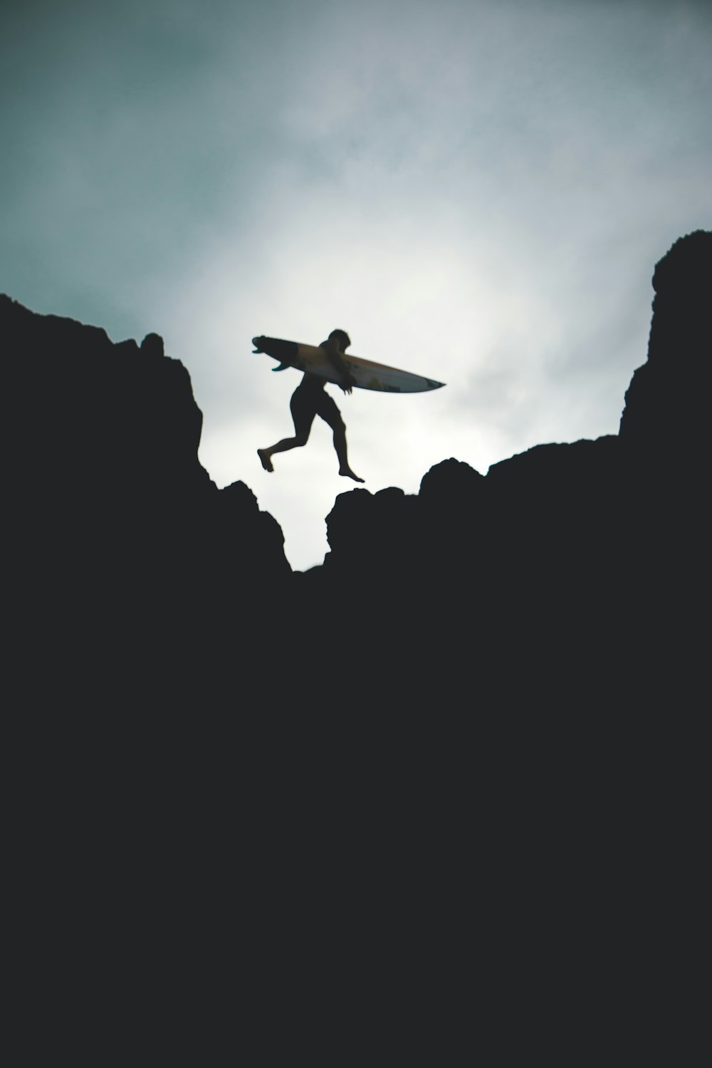 Silhouettenfoto eines Mannes, der auf einem großen Felsen springt und ein Surfbrett hält