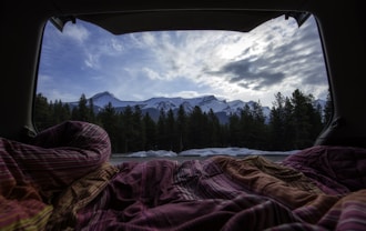 pink, white, and orange blanket inside tent near body of water and forest under gray clouds at daytime