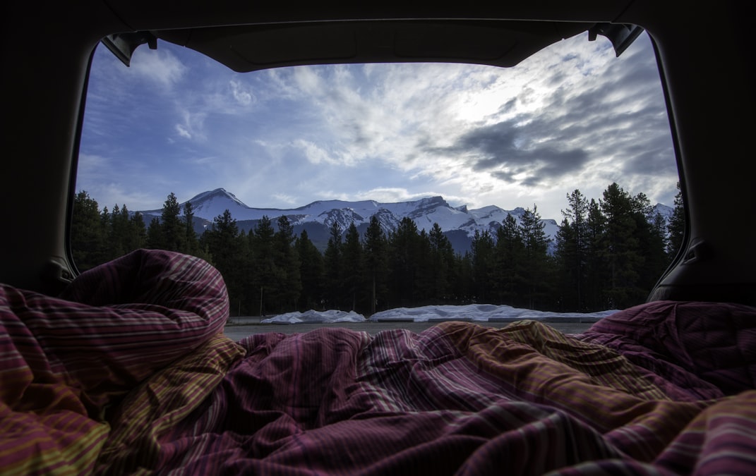View from inside a car at dusk with a sleeping bag in the foreground, snow-capped mountains and pine forest beyond