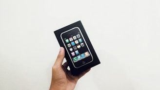 A hand is holding a box containing an older model of a smartphone with a black front and visible app icons. The box is black and features an image of the phone on the front.