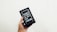 A hand is holding a box containing an older model of a smartphone with a black front and visible app icons. The box is black and features an image of the phone on the front.