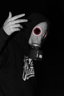 A person wearing a gas mask with a hoodie, holding one hand up against a dark background. The gas mask has prominent circular filters and a distinct, slightly unsettling appearance. The lighting and monochromatic scheme add a dramatic effect.
