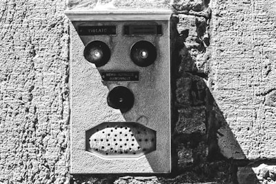 An old intercom system mounted on a textured stone wall. The intercom features three circular buttons and a perforated speaker area. The surface appears worn and aged, adding to the rustic aesthetic of the structure.