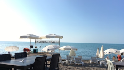 A serene beach setting with several white umbrellas and lounge chairs arranged on the sandy shore. The turquoise sea stretches into the distance under a clear blue sky. A sign reads 'TURKUAZ BEACH' above a small stage area equipped with microphones, likely for live performances. In the foreground, there are dark-colored tables and chairs on a wooden deck.