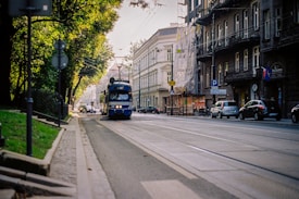 A blue tram is traveling down an urban street lined with parked cars and historic buildings. The street is flanked by trees with dense foliage, casting shadows on the sidewalk. The atmosphere suggests a blend of modernity with classic architecture, and several parked cars are visible.
