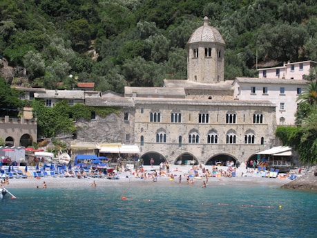 A historic stone building with arched windows and a dome is situated by a beach. The structure is surrounded by lush greenery and trees. Numerous people relax on the beach, enjoying the sun with umbrellas, sunbeds, and towels. The water is calm, and a few small boats are visible near the shore.