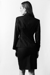 A woman is standing with her back to the camera, wearing a tailored pinstripe suit with a fitted jacket and knee-length skirt. Her hair is long and straight, draped over her shoulder.