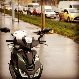 A sleek black scooter with green accents is parked on a wet sidewalk. The scooter features a Waze sticker on the front and a white helmet placed on the seat. In the background, there is a busy street with several cars and vans in traffic. The scene appears to be set on an overcast day, as the pavement glistens with moisture.