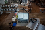 A cozy coffee shop setting with a laptop on a wooden table, surrounded by various items including a cup of coffee, a colorful water bottle, sunglasses, a smartphone, a hard drive, headphones, and an open book. The background shows a counter with stacked coffee products and chairs, adding to the casual ambiance.