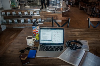 A cozy coffee shop setting with a laptop on a wooden table, surrounded by various items including a cup of coffee, a colorful water bottle, sunglasses, a smartphone, a hard drive, headphones, and an open book. The background shows a counter with stacked coffee products and chairs, adding to the casual ambiance.