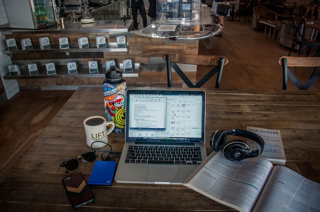 A cozy coffee shop setting with a laptop on a wooden table, surrounded by various items including a cup of coffee, a colorful water bottle, sunglasses, a smartphone, a hard drive, headphones, and an open book. The background shows a counter with stacked coffee products and chairs, adding to the casual ambiance.