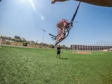 A person holds up a large lacrosse stick on a sunny day in a sports field with green grass and clear skies. Another person stands in the middle of the field near a sports logo, with parking garages and trees in the background.