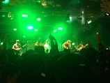 A band performs on stage with bright green lighting creating a vibrant atmosphere. Several musicians are playing various instruments as a large crowd watches, some with their hands raised.