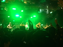 A band performs on stage with bright green lighting creating a vibrant atmosphere. Several musicians are playing various instruments as a large crowd watches, some with their hands raised.