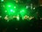 A band performs on stage with bright green lighting creating a vibrant atmosphere. Several musicians are playing various instruments as a large crowd watches, some with their hands raised.