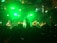 A band performs on stage with bright green lighting creating a vibrant atmosphere. Several musicians are playing various instruments as a large crowd watches, some with their hands raised.